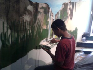 The artist holding a palette while painting the reeds of the mural