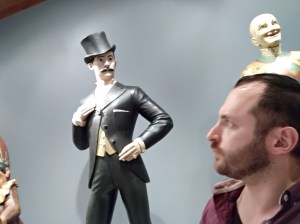Nachliel Selavan pulling a face beside a figurine of a Victorian gentleman in a top hat